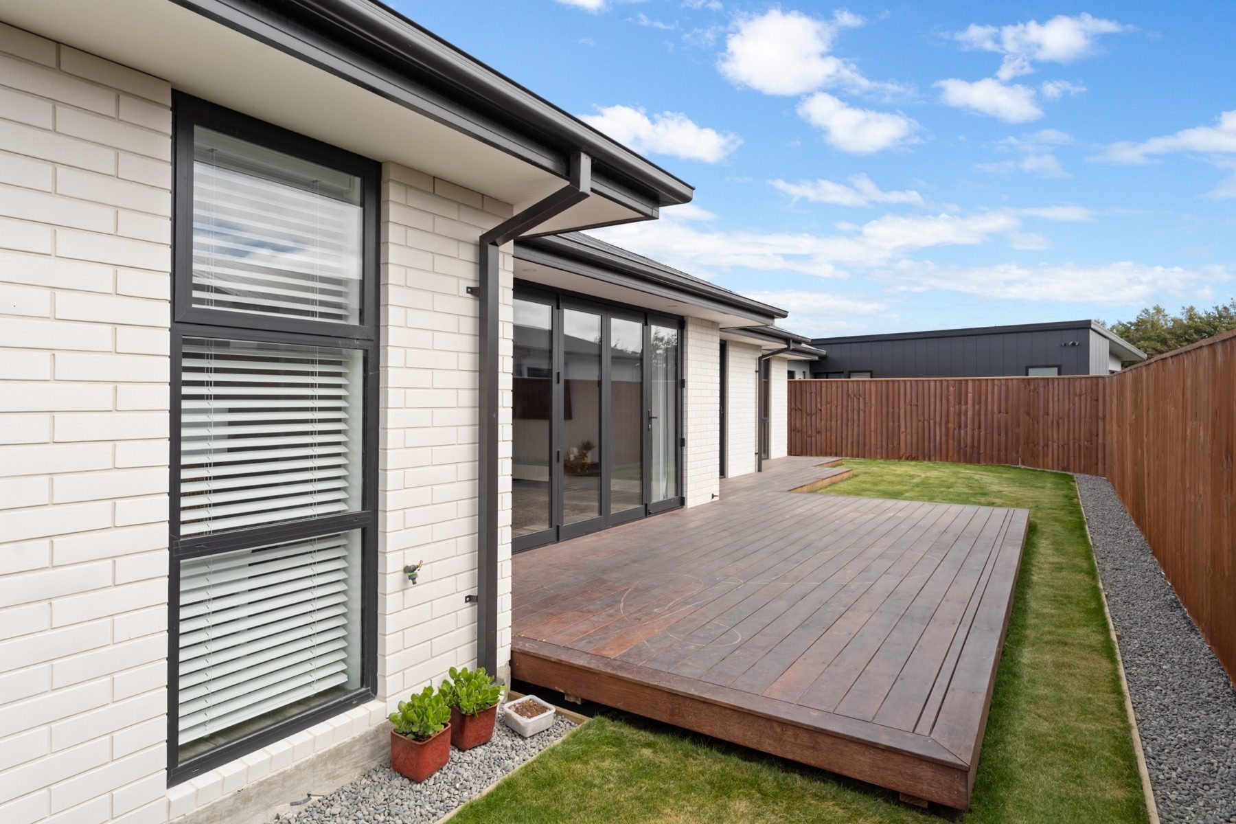 The side courtyard at Milns Park, where the timber deck runs along the pale brick elevation past the bi-fold doors.