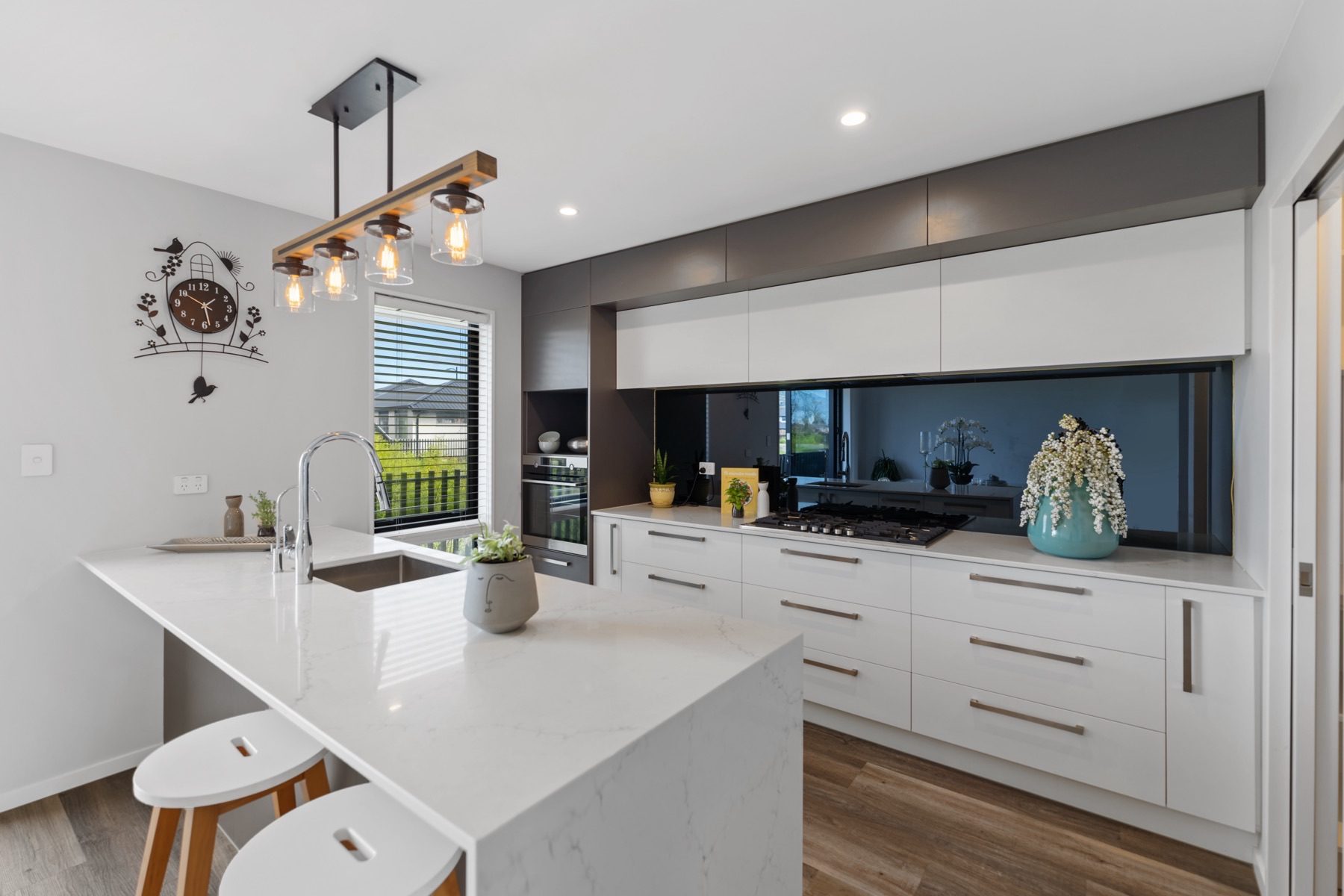 The kitchen from the breakfast bar, showing the waterfall island, a dark glass splashback and dark upper cabinetry above white drawers.