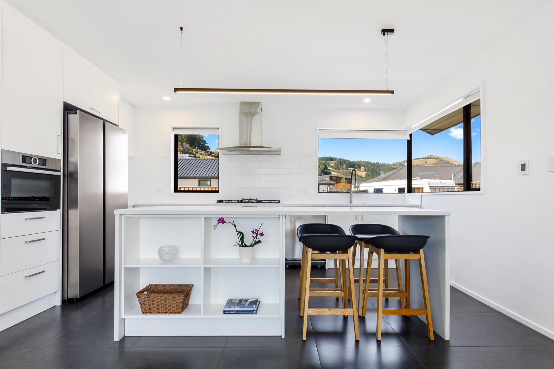 Kitchen with a white island and open end shelving, a stone benchtop, a white tiled splashback and a window framing the hills beyond.