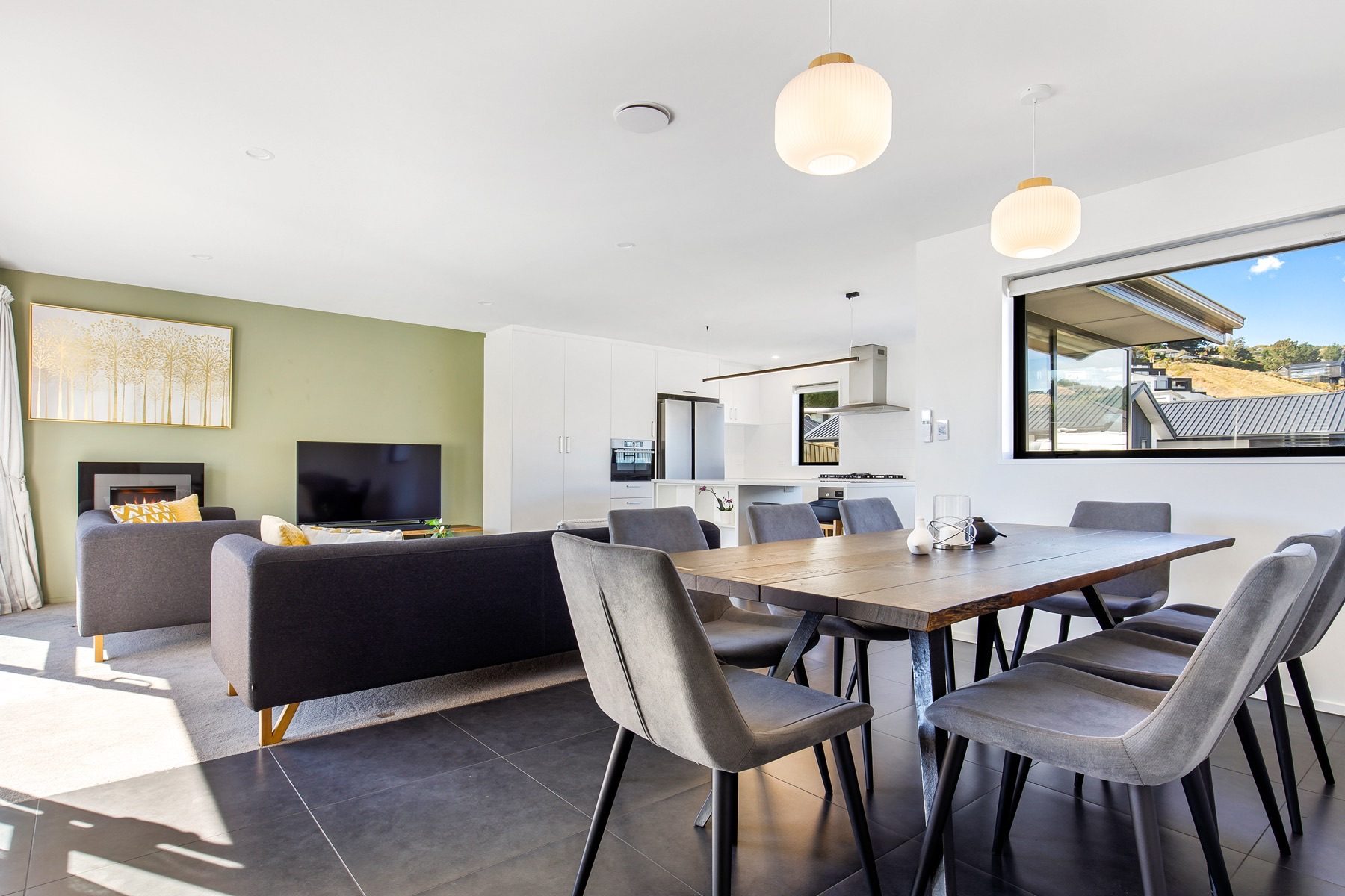 Open-plan dining and living at Old Stone Lane, with a gas fire set into a sage-green wall, ribbed globe pendants over the table and the hills through the far window.