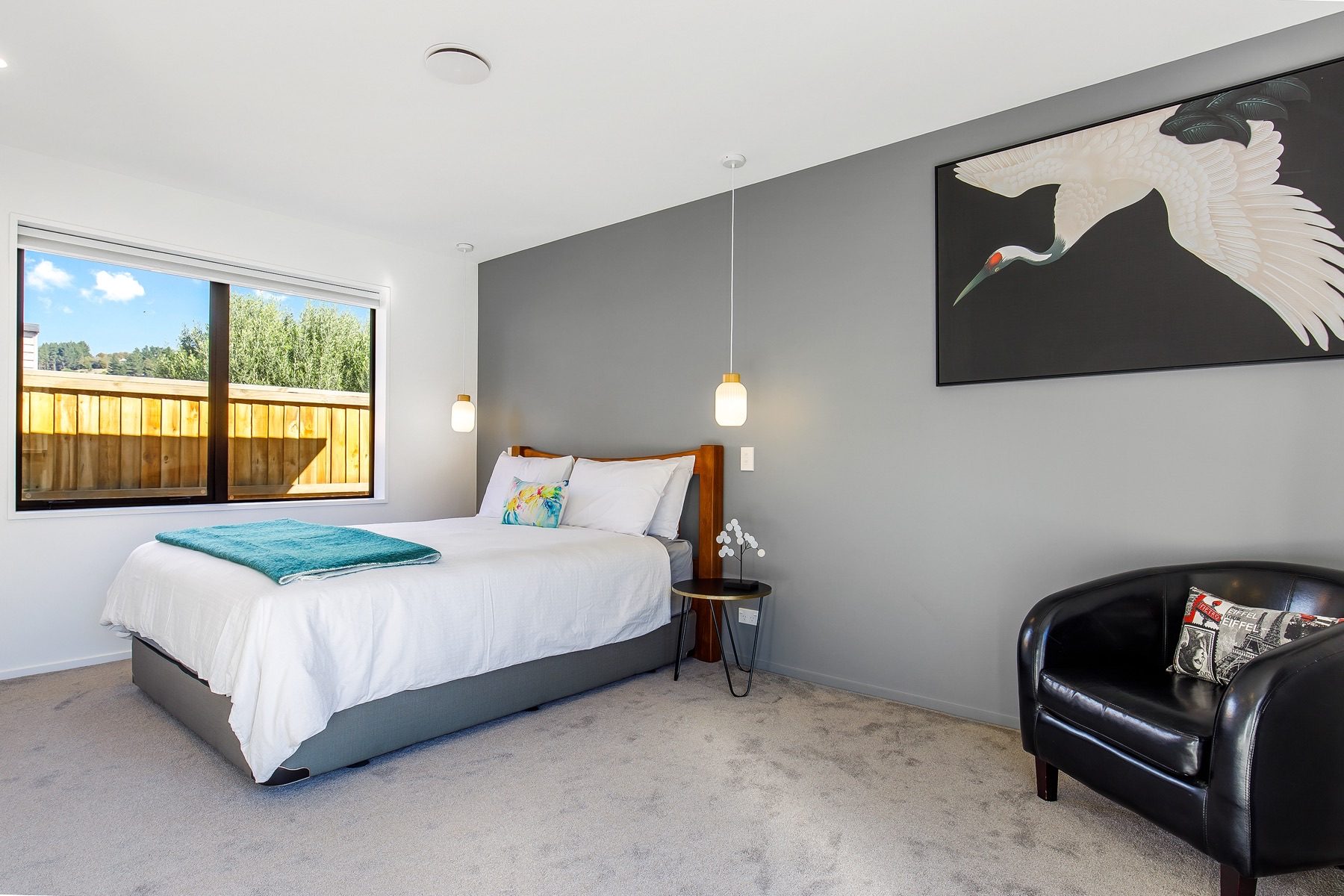 Bedroom with a timber headboard against a grey feature wall, a pendant hung either side of the bed and a window to the garden.