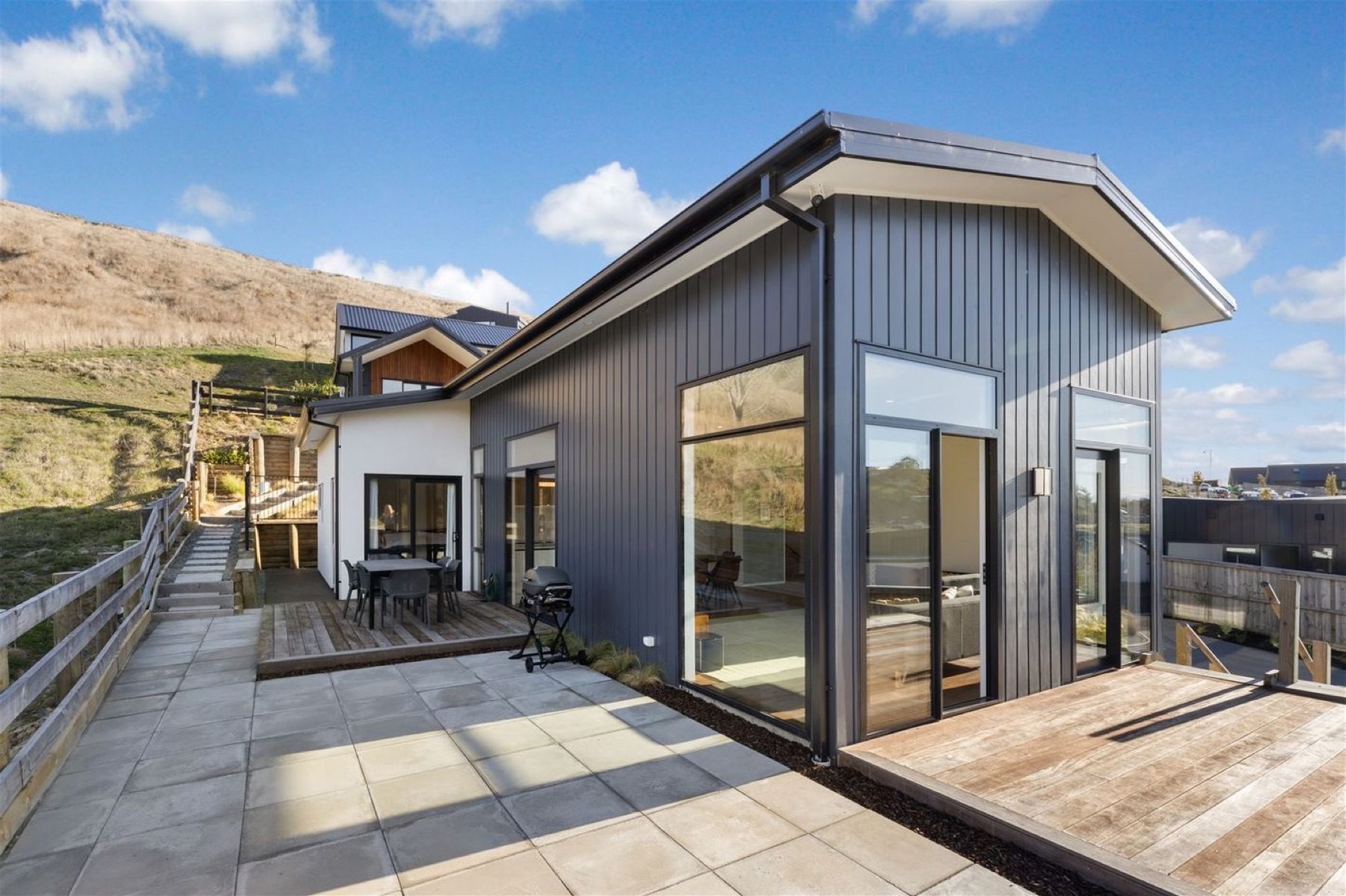 The rear of the Round Hill Rise house, where a paved terrace and timber deck meet glazing that turns the corner, with the hillside paddock rising directly behind the boundary fence.