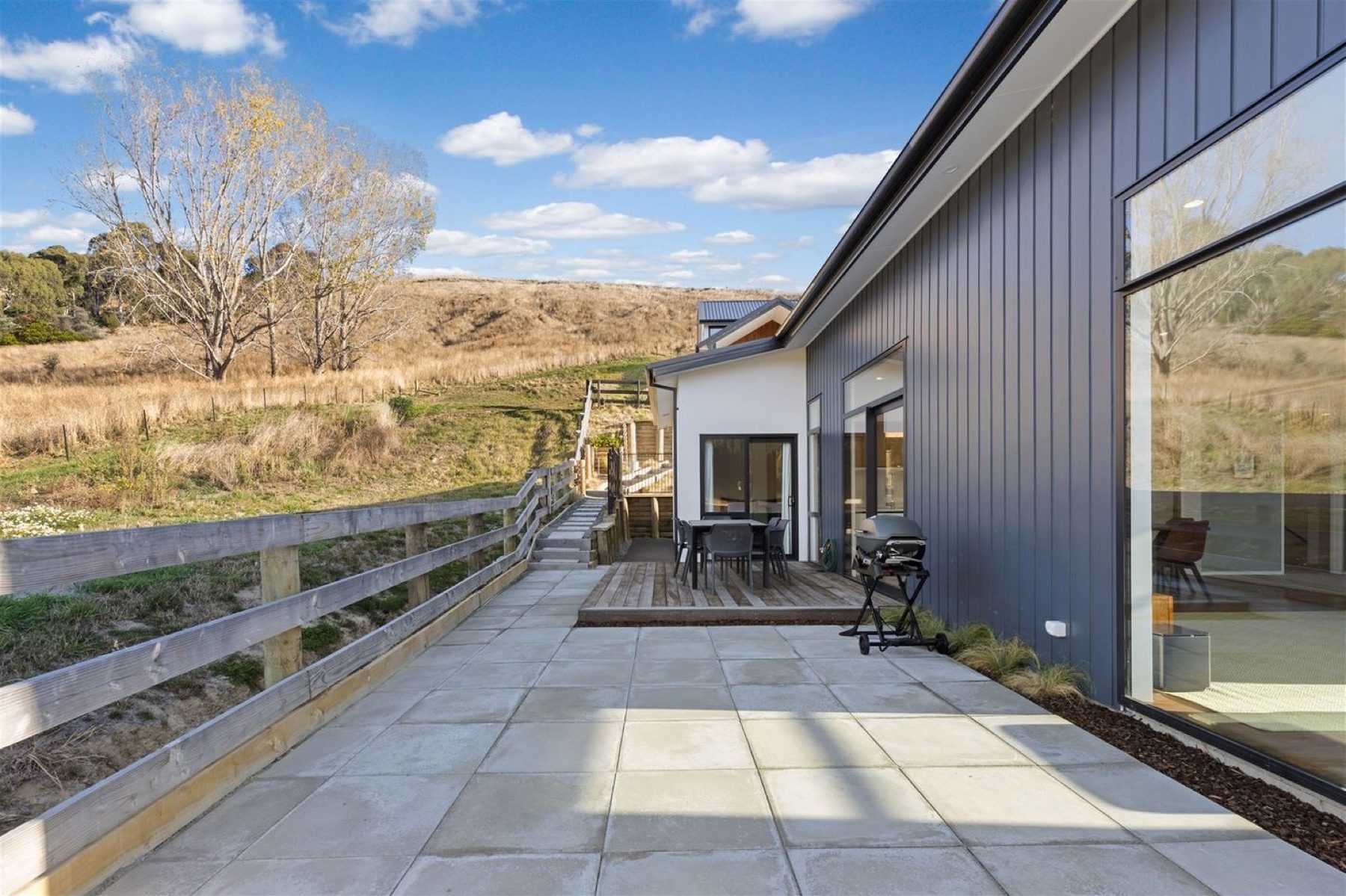 The paved terrace and timber deck running along the dark board-and-batten elevation, with steps climbing the hillside beyond the post-and-rail fence.