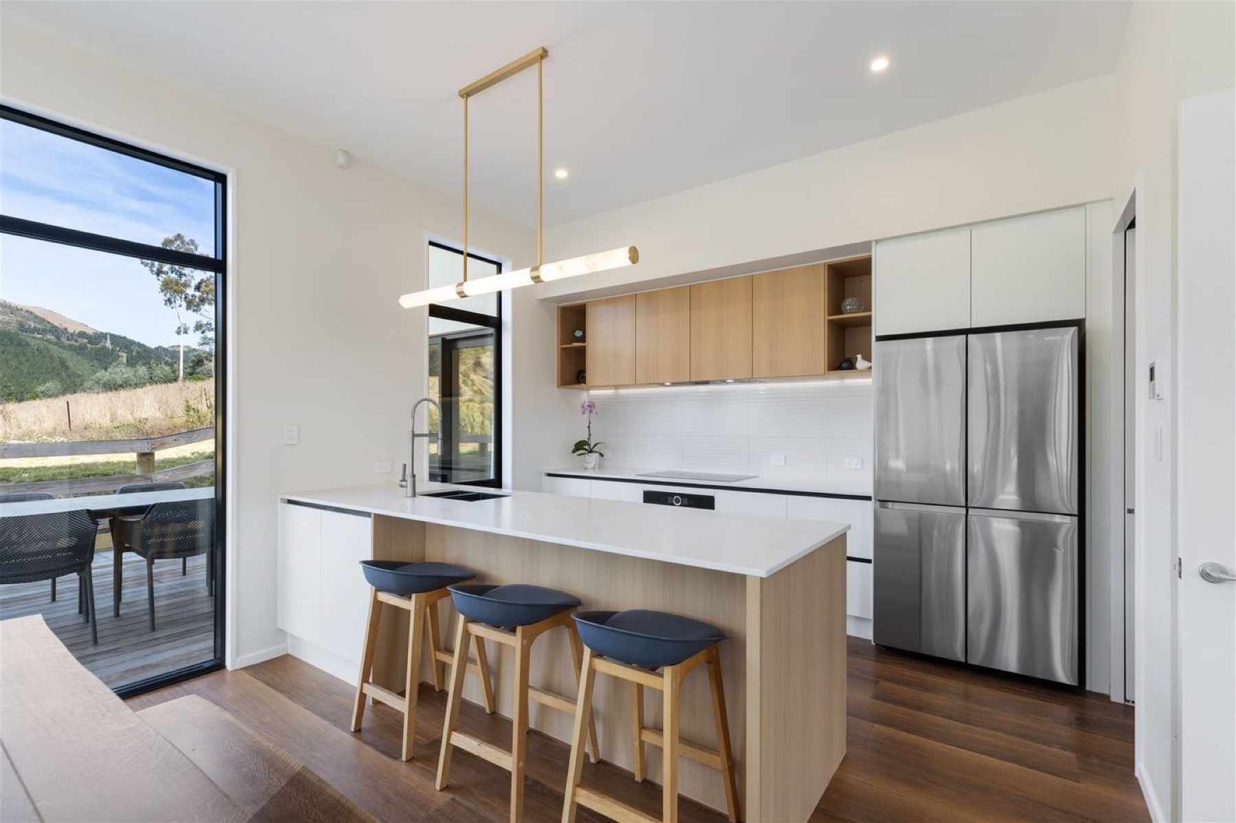 The kitchen looking through to the living area, with a white stone benchtop, oak cabinetry and a ribbed white tile splashback.