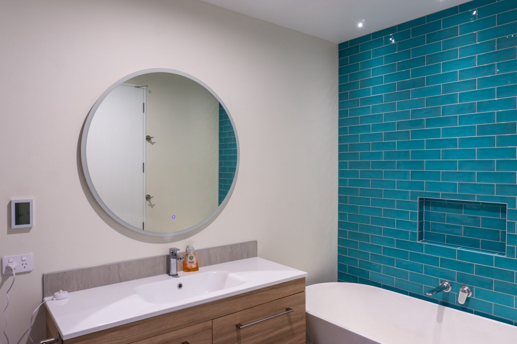 Bathroom with a round mirror, timber vanity and a teal tiled wall.