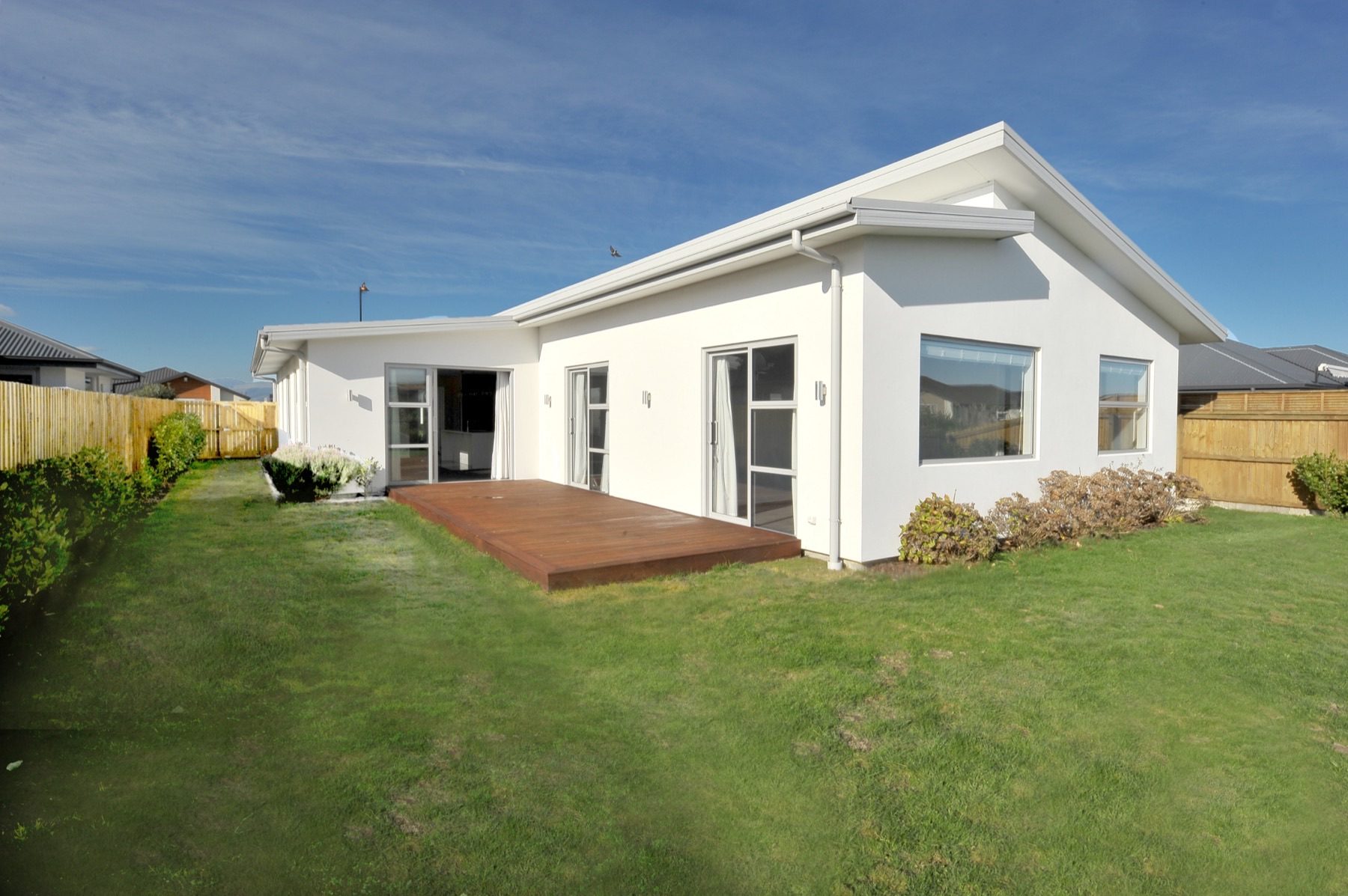 The rear of the Vernon Drive house, where the white render steps down under a low mono-pitch roof to a timber deck and lawn.