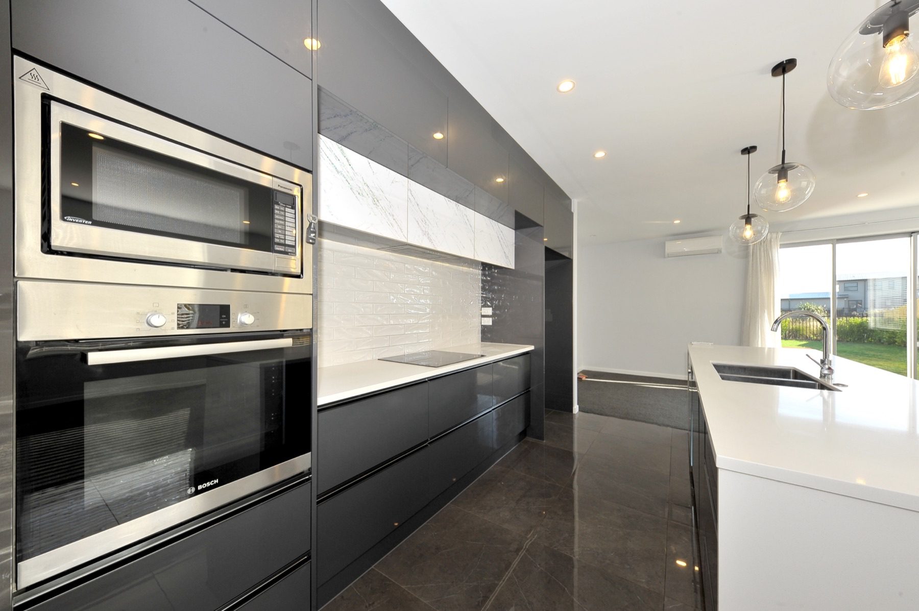 The Vernon Drive kitchen: dark gloss cabinetry, a white stone benchtop and waterfall island, a white tiled splashback and a wall oven.