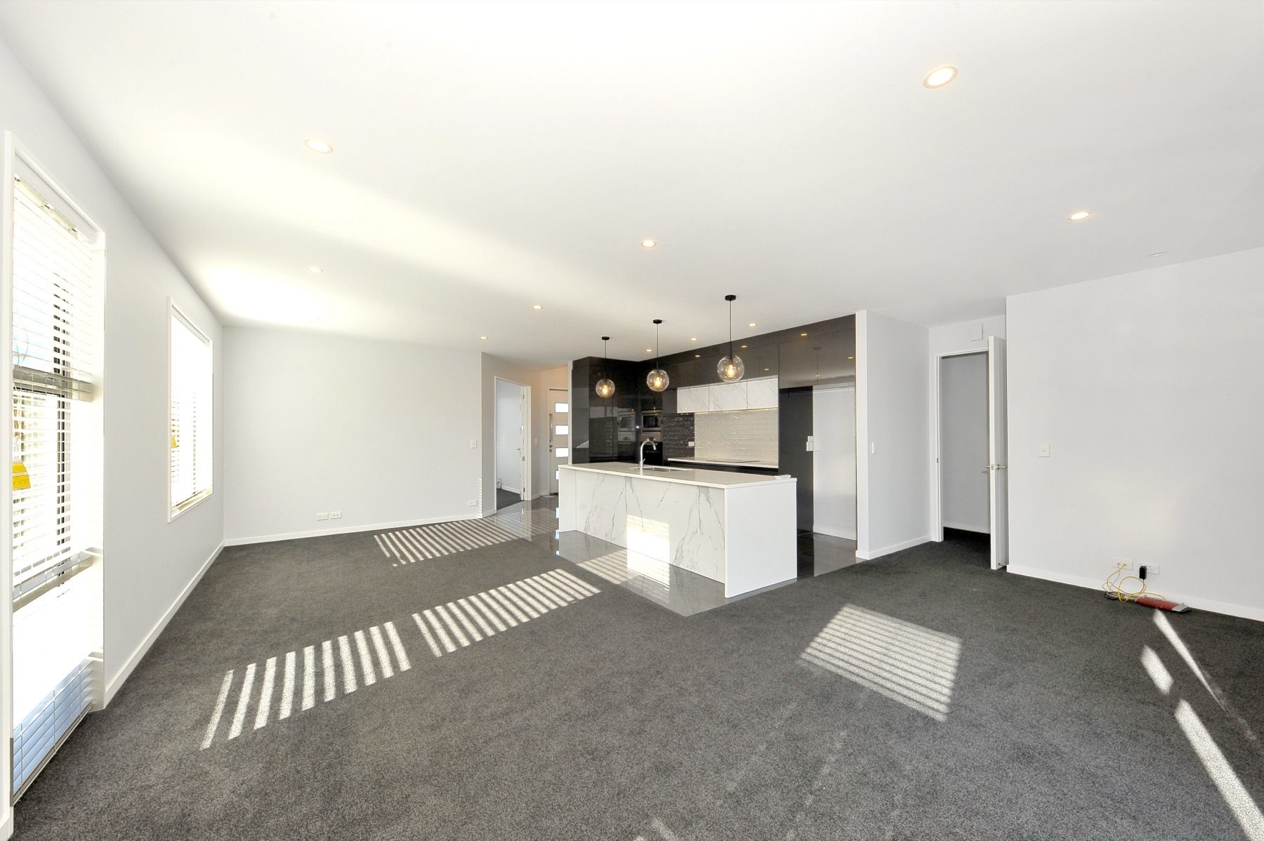 Open-plan living at Vernon Drive, looking across grey carpet to the kitchen's marble-look waterfall island and dark gloss cabinetry under glass globe pendants.