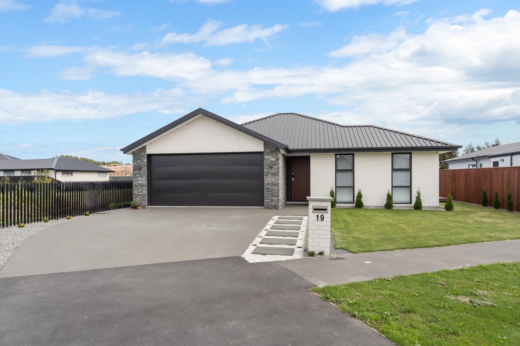 Front elevation of the Milns Park house, with a stone-clad garage, a dark long-run roof and stepping stones across the lawn to the entry.