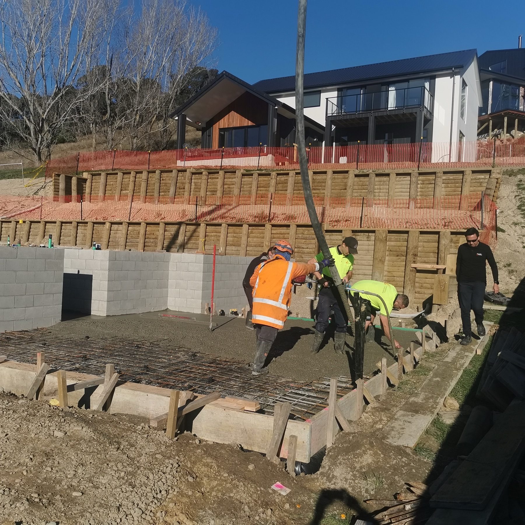 The JSR crew placing concrete into the boxed foundations at Round Hill Rise, Cracroft, with two tiers of timber pole retaining rising behind them.