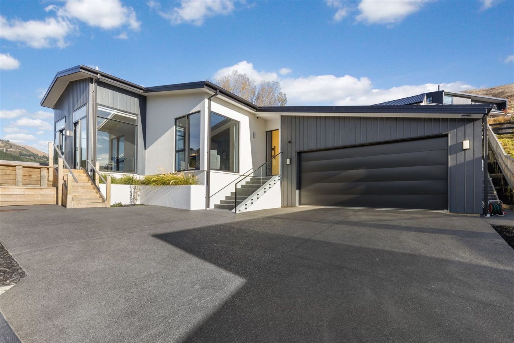 Front elevation of the Round Hill Rise house: dark vertical cladding and white render under opposing skillion roofs, with a yellow front door and concrete steps up from the driveway.